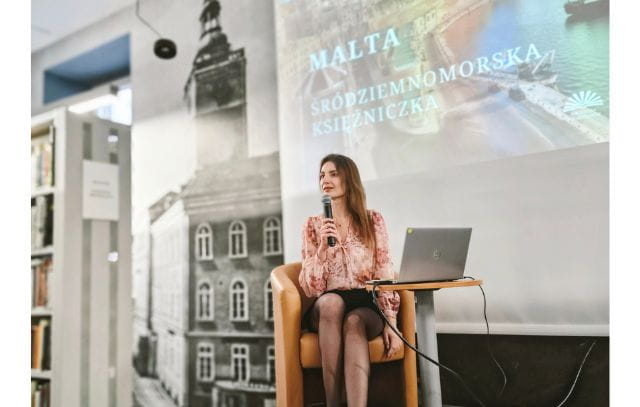 Na zdjęciu prowadząca spotkanie, siedząca przy małym stoliku z komputerem, w ręce trzyma mikrofon. W tle wyświetlona prezentacja o Malcie.