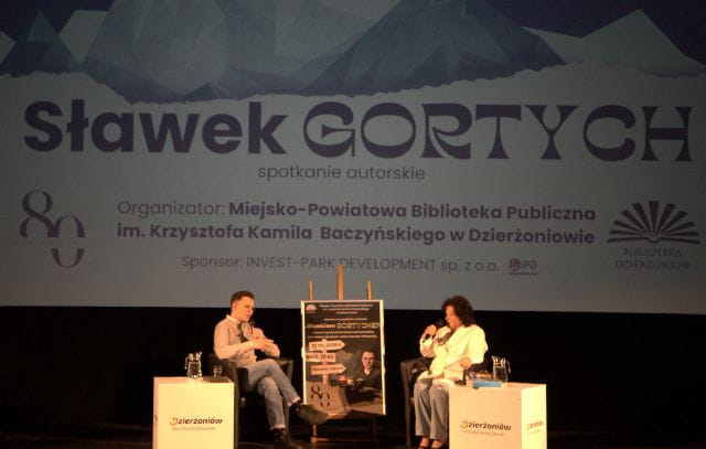 Na scenie w Kinoteatrze Zbyszek siedzą dwie osoby, które ze sobą rozmawiają. W tle widać slajd z górami oraz z napisem Sławek Gortych spotkanie autorskie